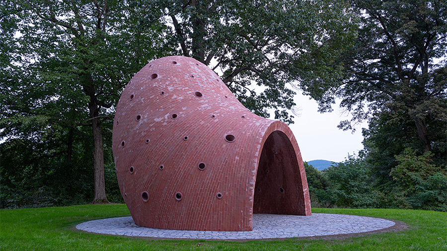 Color photograph of a large inhabitable sculptural structure made from curving red bricks, sited in a forest location