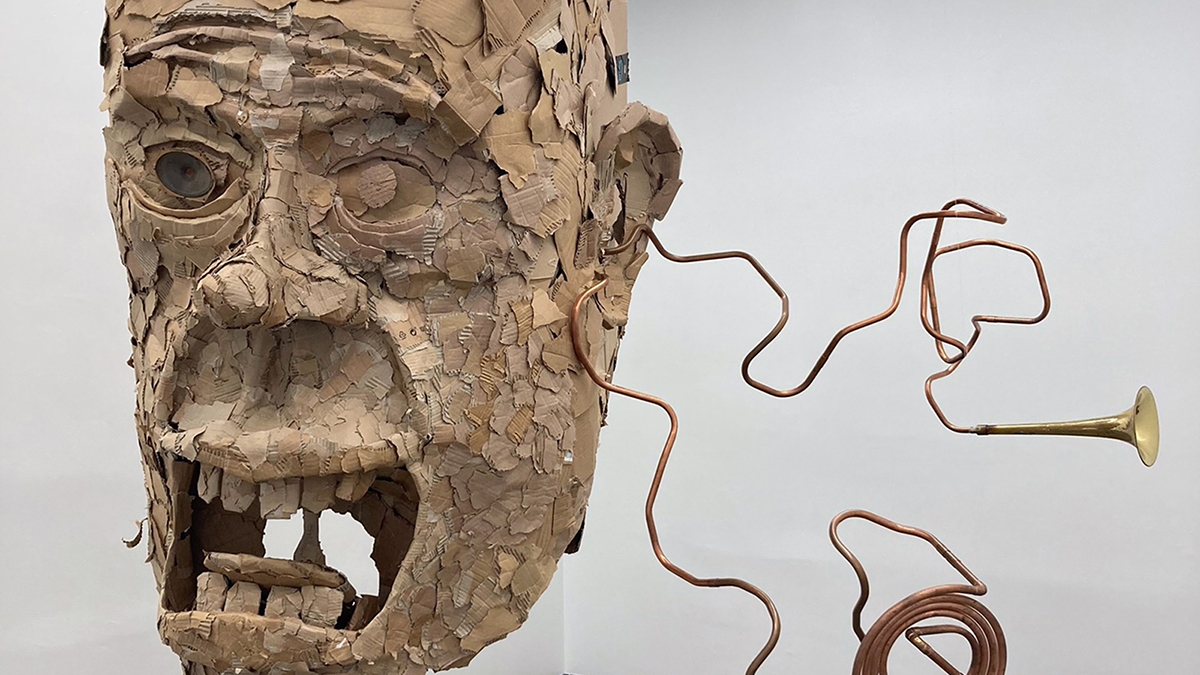 Color photograph of a hanging sculpture that fashions a human head out of cardboard; coming from one ear are twisting tubes of metal that are capped with brass trumpet horns
