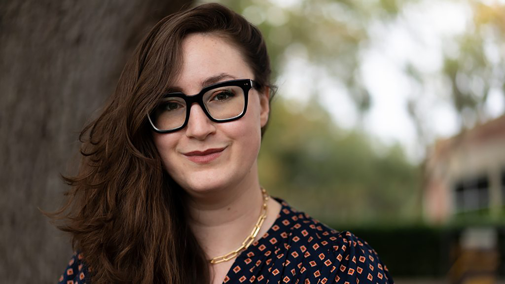 Color photograph of the head of a light skinned woman with brown hair and wearing glasses, standing outdoors next to a tree