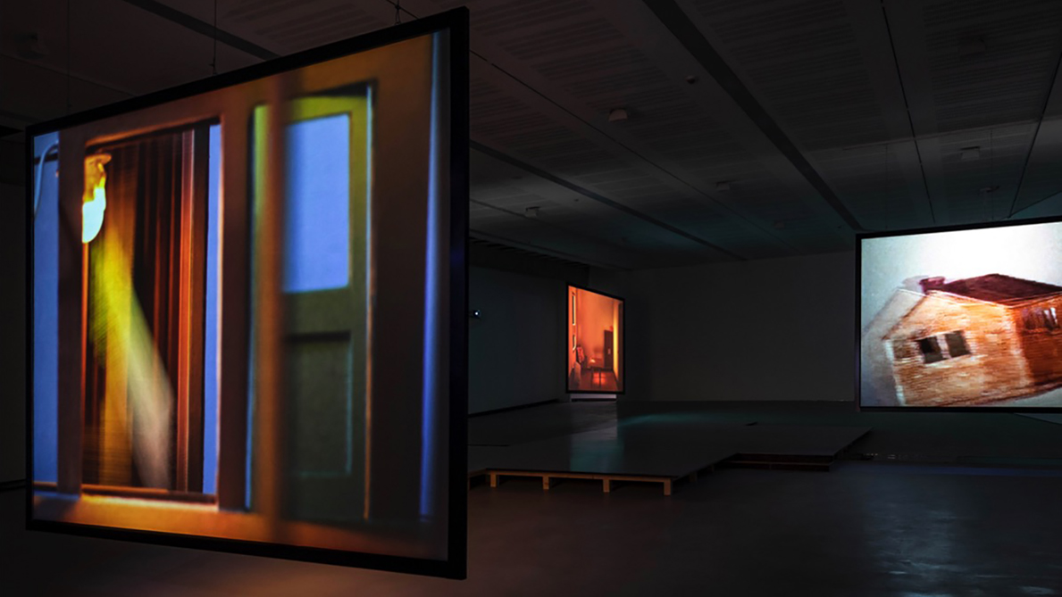 Color photograph of Tomaso De Luca's installation at MAXXII, showing three video screen suspended from the ceiling; on the screen are abstracted imags of house interiors and exteriors