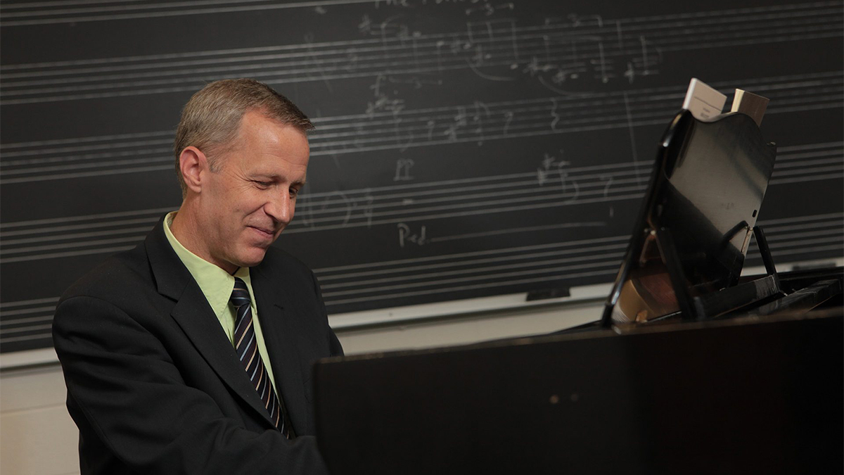 Color photo of a light skinned man sitting at a piano and playing it