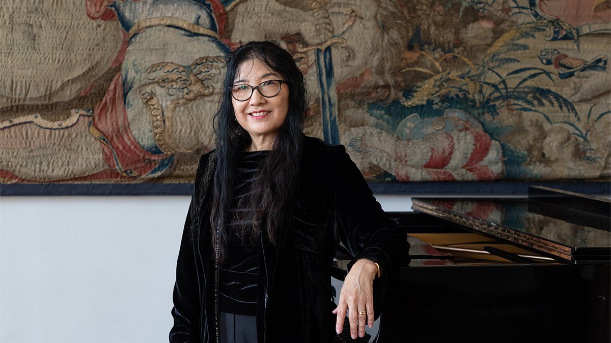 Color photo of an Asian woman wearing glasses and leaning on a piano