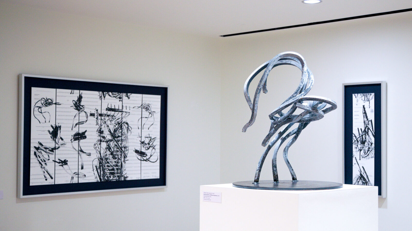 Color photo of an art exhibition with a freestanding twisted steel sculpture and two prints on the wall