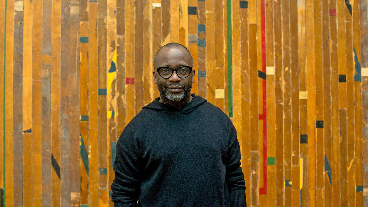 Color portrait of the head and torso of Theaster Gates standing in front of slatted wooden wall