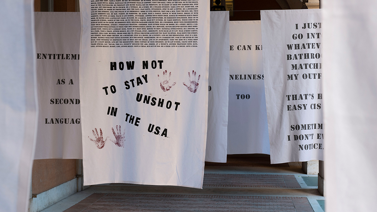 Color photo of white cloth banners hanging in a cortile; on the banners are lines of text and images of handprints, in black