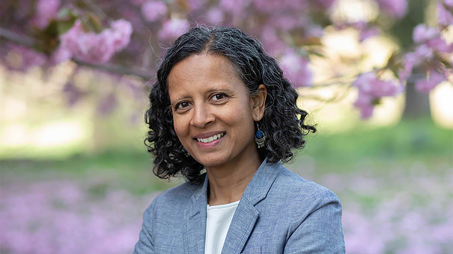 Color photo of the head and shoulders of a brown skinned woman standing in a flower garden, smiling at the camera