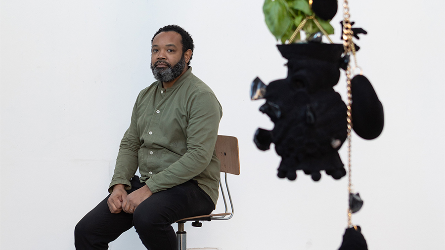 A black man wearing a green shirt and dark pants leans on a chair in a white walled room, with a blurry sculpture made of hanging plants and black velvet hang in the foreground