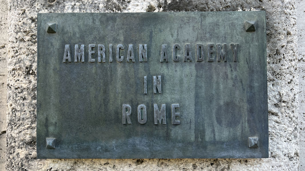 Color photograph of a oxidized steel nameplate on a stone wall; the sign reads American Academy in Rome